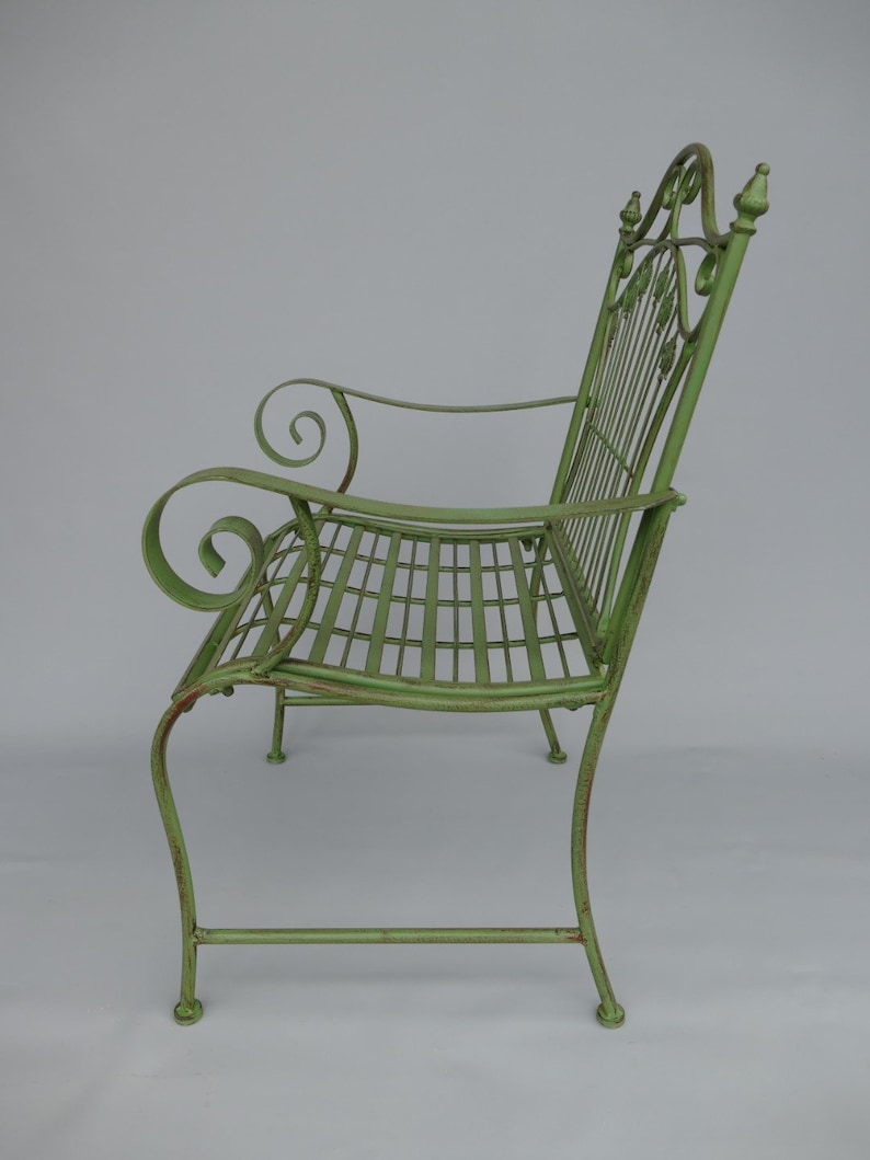 May include: A green metal garden bench with a lattice seat and back. The bench has scrolled armrests and decorative leaf accents on the back. The bench is painted in a distressed, light green color.