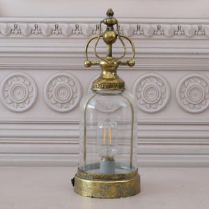 Può includere: Una lampada da tavolo dorata con una cupola di vetro e una parte superiore decorativa. La lampada ha un paralume in vetro trasparente e una base dorata.