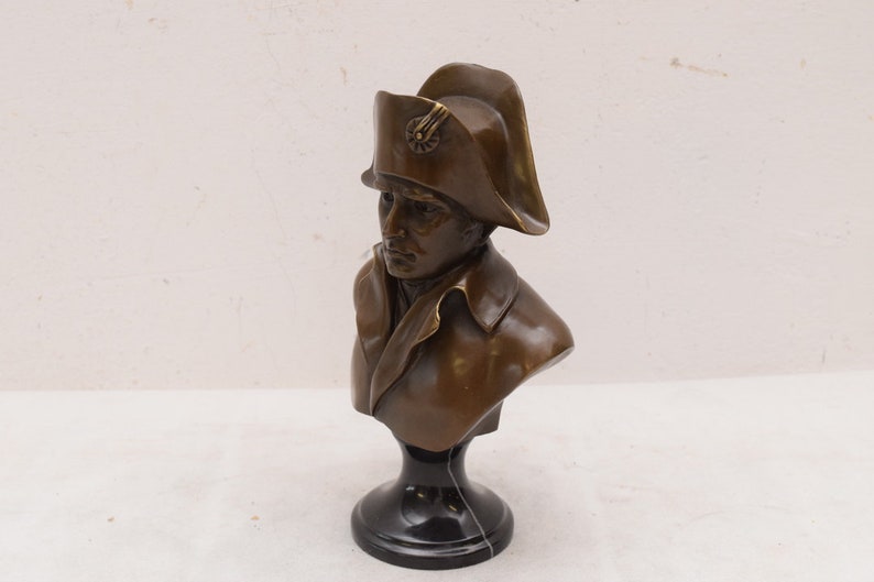 May include: Bronze bust sculpture of a man wearing a bicorne hat. The bust is on a black marble base.