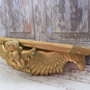 May include: A golden-colored, carved wooden angel shelf. The shelf features a cherub with detailed wings and a rectangular top. The angel is resting its head on its hand. The shelf is painted with gold and green accents.