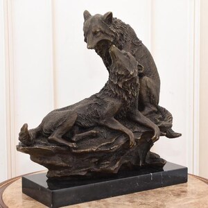 May include: A bronze sculpture of two wolves, one seated and howling, the other standing above it. The sculpture is mounted on a black marble base, showcasing detailed fur and expressive poses. The artwork is a decorative piece.
