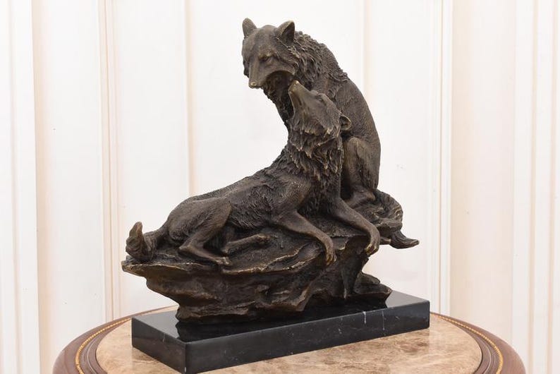 May include: A bronze sculpture of two wolves, one seated and howling, the other standing above it. The sculpture is mounted on a black marble base, showcasing detailed fur and expressive poses. The artwork is a decorative piece.