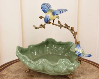 Puede incluir: Un cuenco decorativo de cerámica con un diseño verde ondulado. Dos pájaros de cerámica con colores azul y amarillo se posan en una rama de color bronce que se arquea sobre el cuenco. El cuenco tiene patas de color bronce.