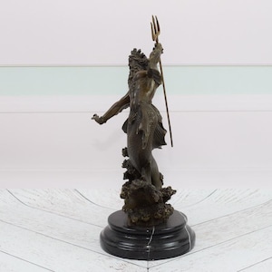 Puede incluir: Estatua de bronce de una figura m&iacute;tica, posiblemente Neptuno, sosteniendo un tridente. La figura est&aacute; posada con un brazo extendido y se alza sobre una base texturizada. La estatua tiene una p&aacute;tina marr&oacute;n oscuro y est&aacute; montada sobre una base de m&aacute;rmol negro.