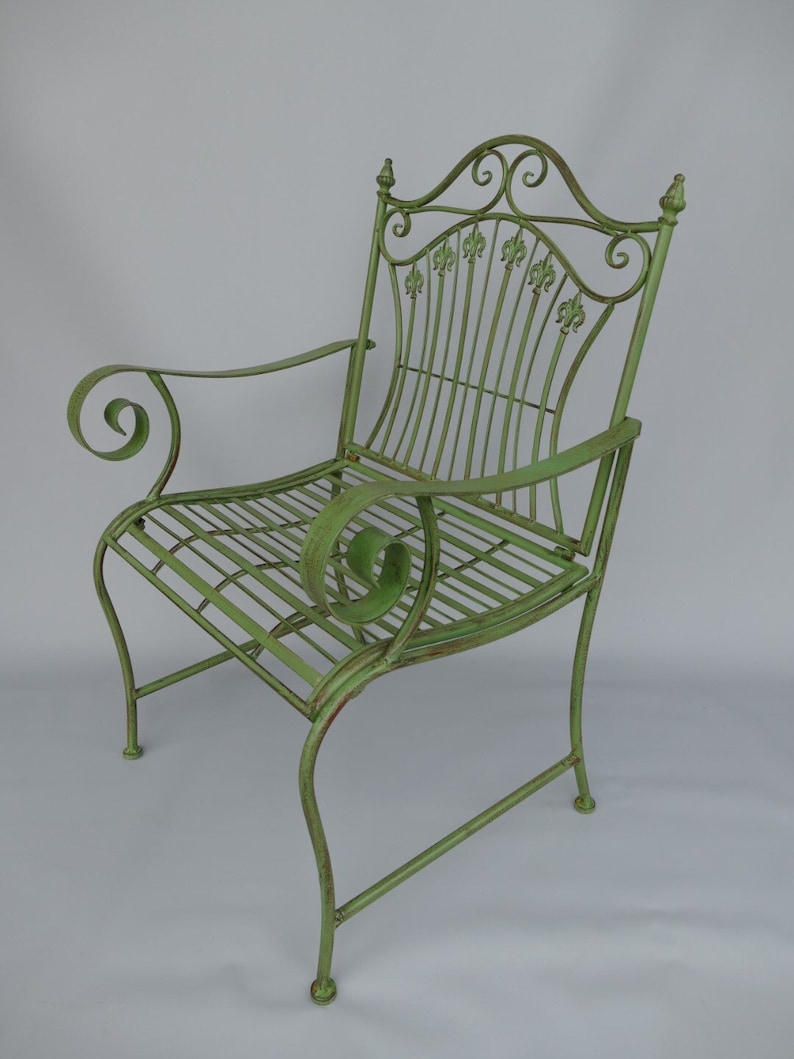 May include: A distressed green metal garden chair with a decorative back featuring fleur-de-lis accents. The chair has curved armrests and a slatted seat. The overall design is ornate and suitable for outdoor use.