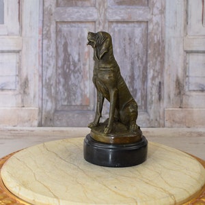 Op de afbeelding: Bronzen beeld van een hond die op een zwarte marmeren voet zit. De hond kijkt omhoog en heeft een lange snuit. Het beeld staat op een ronde, witte marmeren tafel met gouden rand.