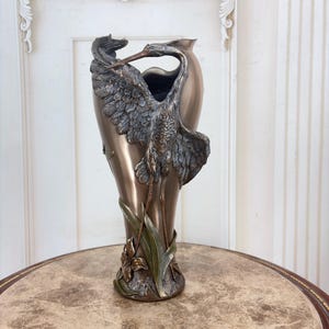 Könnte beinhalten: Eine hohe, metallisch bronzefarbene Vase mit einem skulptierten Reiher, dessen ausgebreitete Flügel den oberen Teil umschließen. Der Sockel ist mit reliefartigen grünen Blättern und einer bronzenen Blume verziert, was einen dreidimensionalen Effekt erzeugt.