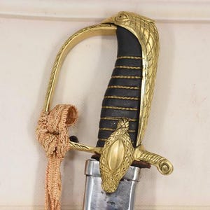 Puede incluir: Primer plano de una espada decorativa con una empuñadura dorada en forma de cabeza de águila y un mango negro. La espada presenta una guarda dorada y una borla decorativa. La hoja es plateada.