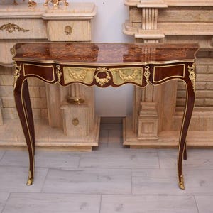 Puede incluir: Una consola de color caoba con acabado brillante, detalles dorados y ornamentados. La mesa tiene patas curvas y un faldón decorativo. La mesa mide aproximadamente 76 cm de largo.