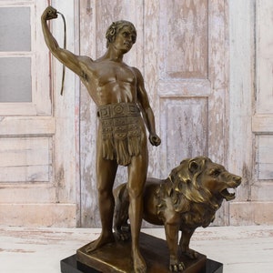 May include: Bronze sculpture of a man holding a whip standing next to a lion. The sculpture is on a black marble base.