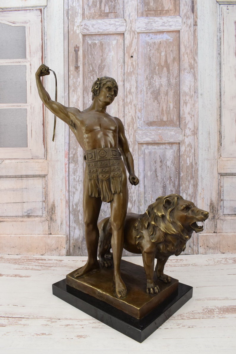 May include: Bronze sculpture of a man holding a whip standing next to a lion. The sculpture is on a black marble base.