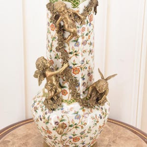 May include: A white porcelain vase with a floral pattern and bronze cherub accents. The vase has a gold rim and base, with cherubs climbing floral garlands. The vase is approximately 18 inches tall.
