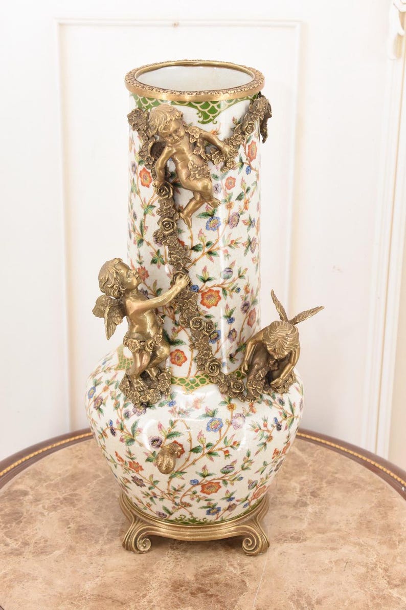 May include: A white porcelain vase with a floral pattern and bronze cherub accents. The vase has a gold rim and base, with cherubs climbing floral garlands. The vase is approximately 18 inches tall.