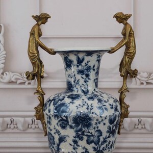 May include: A white porcelain vase with a blue and white floral pattern. The vase has two gold handles shaped like women holding the vase.
