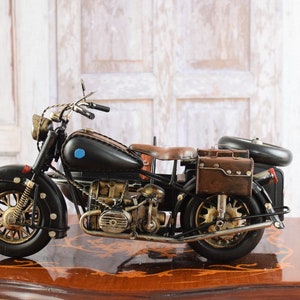 May include: A detailed black and brown vintage motorcycle model with gold-colored accents. The model features a leather seat, saddlebags, and a classic design. The motorcycle is displayed on a wooden surface.