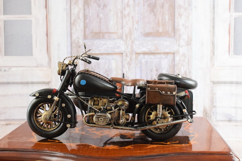 May include: A detailed black and brown vintage motorcycle model with gold-colored accents. The model features a leather seat, saddlebags, and a classic design. The motorcycle is displayed on a wooden surface.