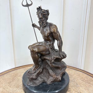 May include: A bronze sculpture of a seated male figure, holding a trident. The figure is atop a rock formation, mounted on a black marble base. The sculpture has a detailed, aged appearance, with a crown and beard.