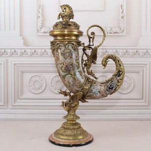 May include: A large, ornate, gold-colored, decorative vase with a white and gold design. The vase is shaped like a horn and has a lid with a finial. The vase is on a pedestal with a marble base.
