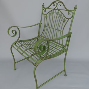 May include: A distressed green metal garden chair with a fleur-de-lis design on the backrest. The chair features curved armrests and a slatted seat. The metal has a weathered appearance, suggesting outdoor use. The chair is a classic piece of outdoor furniture.