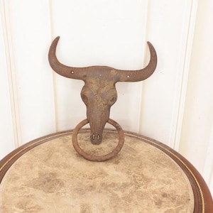 May include: A weathered metal bull skull wall decoration with a large ring below the skull. The skull is mounted above a round table with a light brown surface and a dark brown border. The piece has a vintage, Western aesthetic.