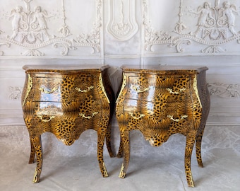 May include: A pair of matching bedside tables featuring a glossy leopard print finish. Each table has two drawers with gold-toned metal handles and curved legs. The front edges of the tables are accented with decorative gold-toned metal trim.