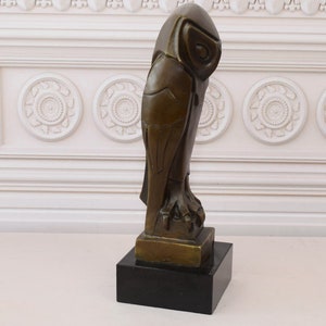 May include: A bronze sculpture of an owl with geometric details, standing on a black base. The owl is facing to the right, with its head turned to the left.