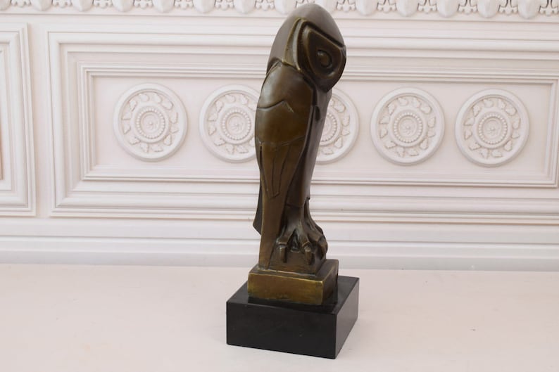 May include: A bronze sculpture of an owl with geometric details, standing on a black base. The owl is facing to the right, with its head turned to the left.