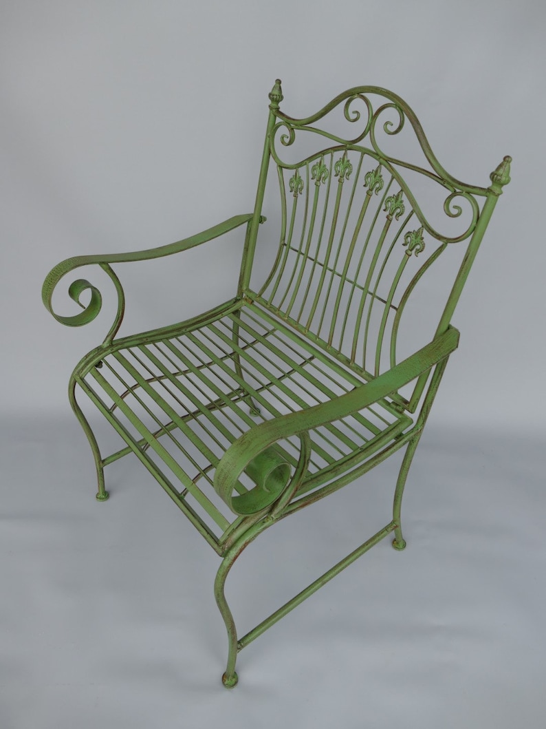 May include: A green metal garden chair with a scrolled back and armrests. The chair features a slatted seat and back, with decorative fleur-de-lis accents. The chair's dimensions are approximately 36 inches high, 24 inches wide, and 24 inches deep.