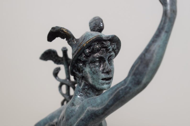 May include: A bronze statue of a figure with a hat and raised arm. The statue has a weathered, verdigris finish, with details like a winged staff and a hat with a gold trim. The sculpture is a classical art piece.