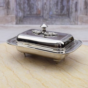 May include: A silver butter dish with a lid and a decorative floral design. The dish has three legs and is sitting on a light brown surface.
