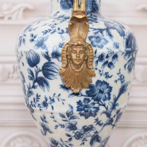 May include: A white porcelain vase with a blue and white floral pattern. The vase has a gold-colored handle with a decorative face.