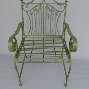 May include: A light green metal garden chair with a decorative back featuring fleur-de-lis accents. The chair has curved armrests and a slatted seat. The overall design is ornate and vintage-inspired, suitable for outdoor use.