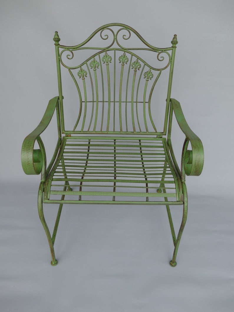 May include: A light green metal garden chair with a decorative back featuring fleur-de-lis accents. The chair has curved armrests and a slatted seat. The overall design is ornate and vintage-inspired, suitable for outdoor use.