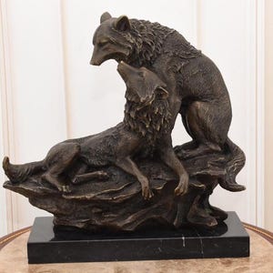 May include: A bronze sculpture of two wolves. One wolf is lying down, looking up at the other wolf, which is standing above it. The sculpture is mounted on a black rectangular base. The wolves are detailed with textured fur.