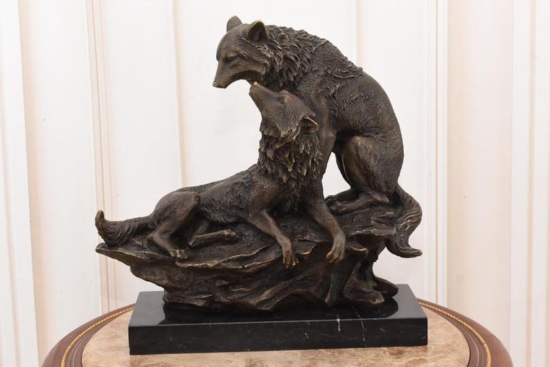 May include: A bronze sculpture of two wolves. One wolf is lying down, looking up at the other wolf, which is standing above it. The sculpture is mounted on a black rectangular base. The wolves are detailed with textured fur.