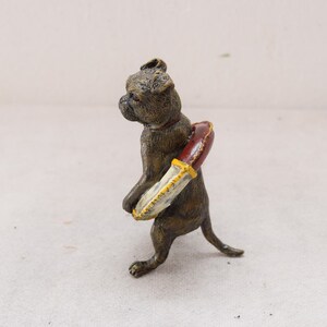 May include: A bronze-coloured figurine of a dog carrying a red and cream-coloured object. The dog is a Boxer breed, with a short snout and upright ears. The object appears to be a decorative item, possibly a knife or a decorative element.