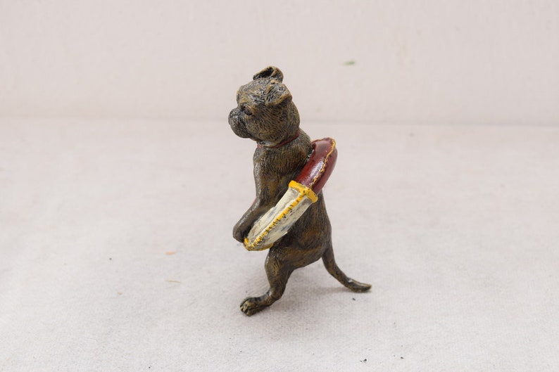 May include: A bronze-coloured figurine of a dog carrying a red and cream-coloured object. The dog is a Boxer breed, with a short snout and upright ears. The object appears to be a decorative item, possibly a knife or a decorative element.
