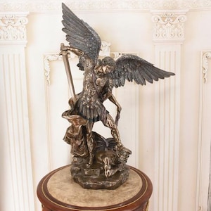 May include: A bronze-toned statue of an archangel with large, detailed wings, wearing Roman-style armor and holding a sword raised high. The figure stands over a defeated, crouching creature on a rocky base, capturing a dynamic, heroic pose.
