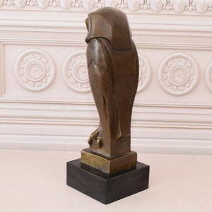 May include: A bronze sculpture of an owl with a black marble base. The owl is standing on a base and is facing to the right. The owl is stylized and has a geometric design.