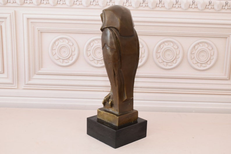 May include: A bronze sculpture of an owl with a black marble base. The owl is standing on a base and is facing to the right. The owl is stylized and has a geometric design.