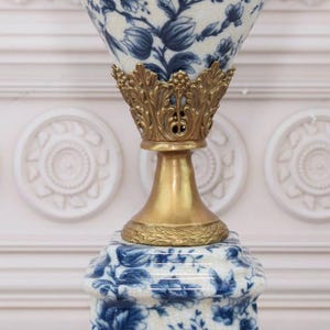 May include: A white ceramic lamp base with a blue floral pattern and a gold metal top. The lamp base is in the shape of a vase with a wide mouth and a narrow neck.