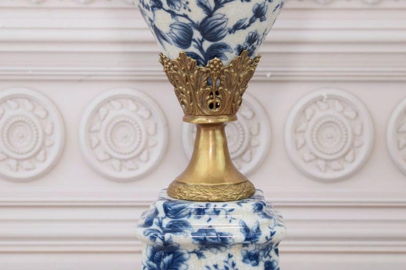 May include: A white ceramic lamp base with a blue floral pattern and a gold metal top. The lamp base is in the shape of a vase with a wide mouth and a narrow neck.
