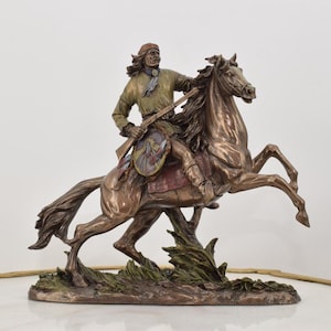 Puede incluir: Una escultura de bronce de un guerrero nativo americano montando un caballo. El guerrero sostiene un rifle y lleva un tocado de plumas. El caballo se levanta sobre sus patas traseras.