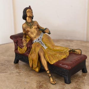 May include: A gold-toned statue of a reclining figure on a burgundy chaise lounge. The figure wears a yellow gown and a decorative headpiece. The chaise lounge has a black base and is placed on a marble surface.