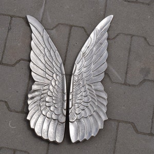 May include: A pair of silver angel wings with a detailed feathered design. The wings are made of metal and have a smooth, polished finish.