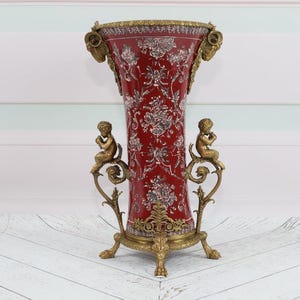 Può includere: Un vaso in porcellana rosso e bianco con un motivo floreale e dettagli dorati. Il vaso è su un piedistallo in metallo dorato con figure di cherubini su ogni lato.