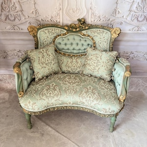 May include: A light green and gold loveseat featuring a tufted backrest, ornate carved wooden frame with gold accents, and two matching square throw pillows. The upholstery displays a detailed damask pattern, and a small cherub figure sits atop the crest.
