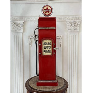 May include: A vintage-style red metal petrol pump replica featuring a circular top sign with a red star and the word GASOLINE. The front display panel shows the text THIS SALE, GALLON, and TOTAL AMOUNT. A black hose is attached to the side.