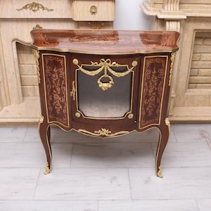 Puede incluir: Pequeño mueble antiguo de madera, acabado en marrón oscuro con detalles dorados. El mueble presenta diseños florales y de volutas ornamentados, una puerta de cristal y patas curvas. La parte superior también está decorada con adornos dorados. Mide unos 80 cm de alto.