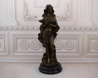 May include: A bronze sculpture of a woman holding a bird in her hands. The sculpture is standing on a black base and is set against a white background.